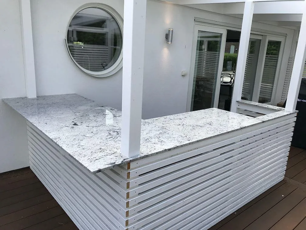 Køkkenbordplade i hvid Antique White-granit, poleret, monteret som vinklad utomhusbänk på vita spjälstommar på ett trädäck i Dösjebro.