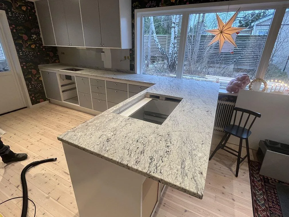Mat køkkenbordplade i lys Antique White-granit på køkkenø og bänkrad vid vindue i Segeltorp.