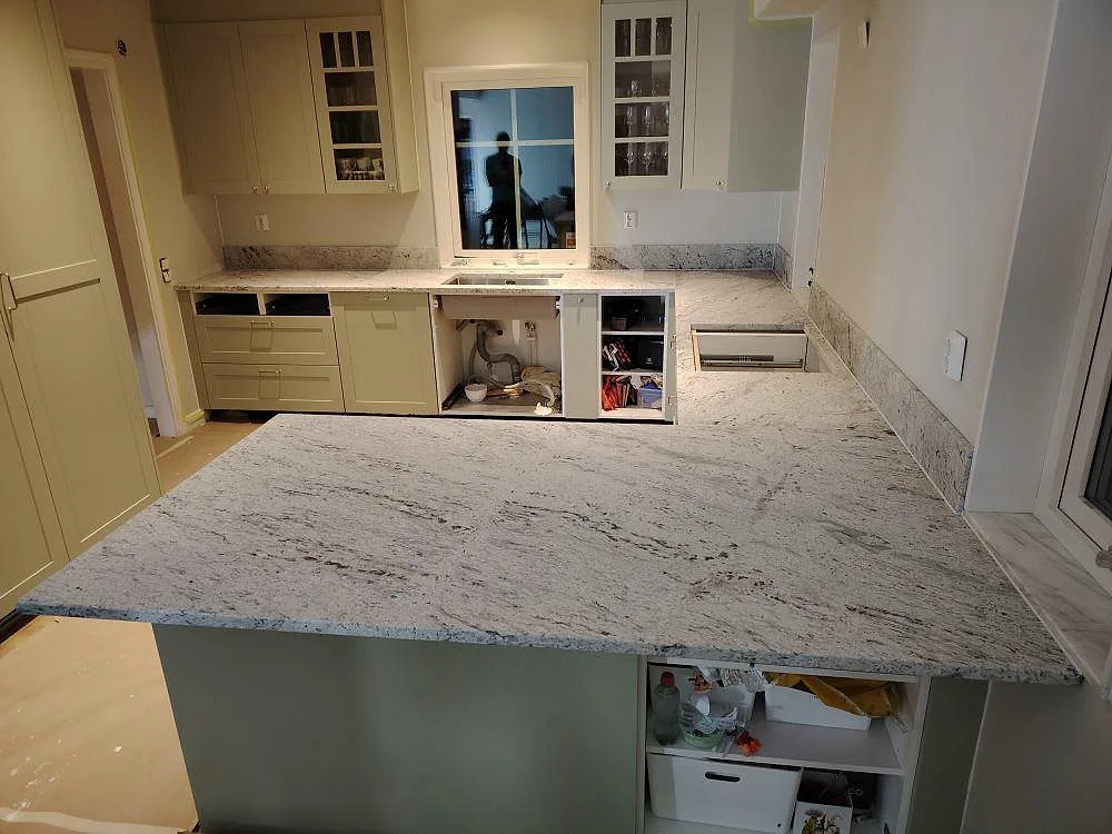 Køkkenbordplade i mat Antique White granit med stor halvö, lys åretegning og låg stänkkant i ett køkken i Västerås.