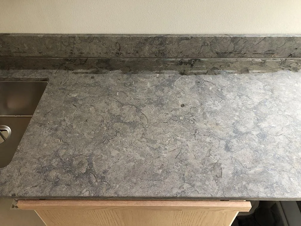 Køkkenbordplade i grå Azul Valverde-granit, mat overflade, längs en köksvägg med lyse eklaminerade överskåp, rostfri köksfläkt og underlimmad vask i ett køkken i Malmö.