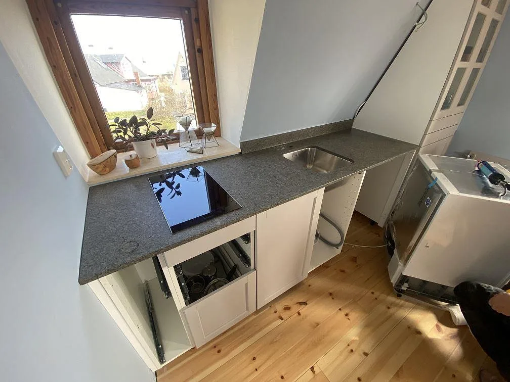 Køkkenbordplade i grå granit Bohus Grå, mat finish, längs fönstret i ett litet køkken under snedtak med underlimmad rostfri vask og induktionshäll i Brantevik.