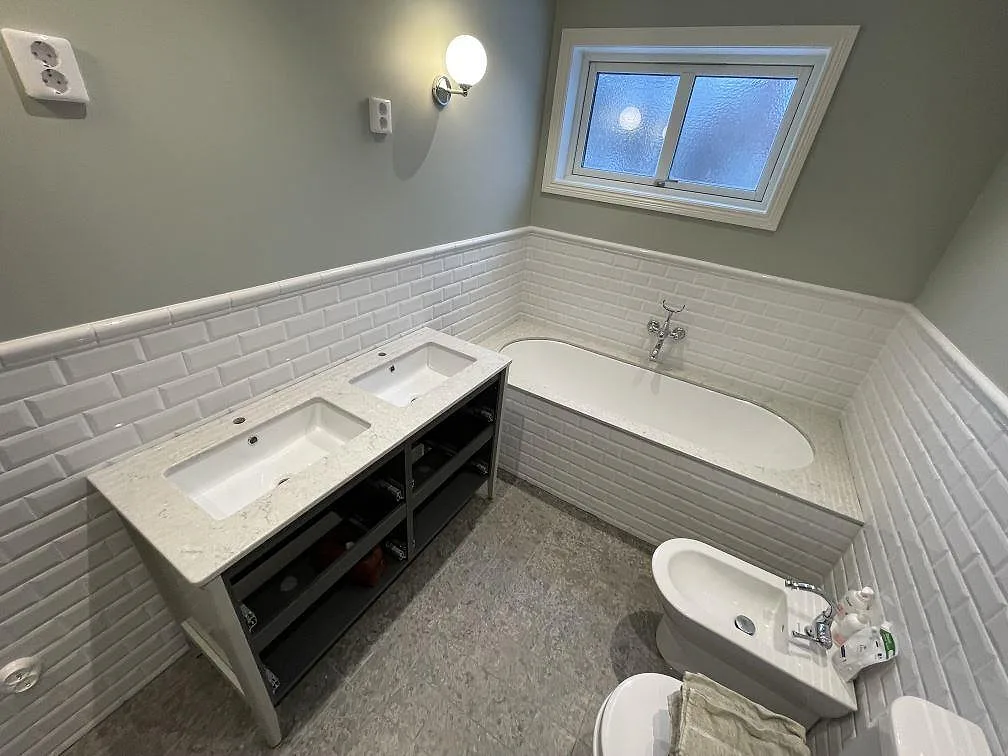 Bordplade i hvid Caesarstone Bianco Drift, poleret, som tvättställsskiva med två underlimmade handfat på en mørk kommod og matchande stenram rundt om badkaret i ett badeværelse i Enskede.