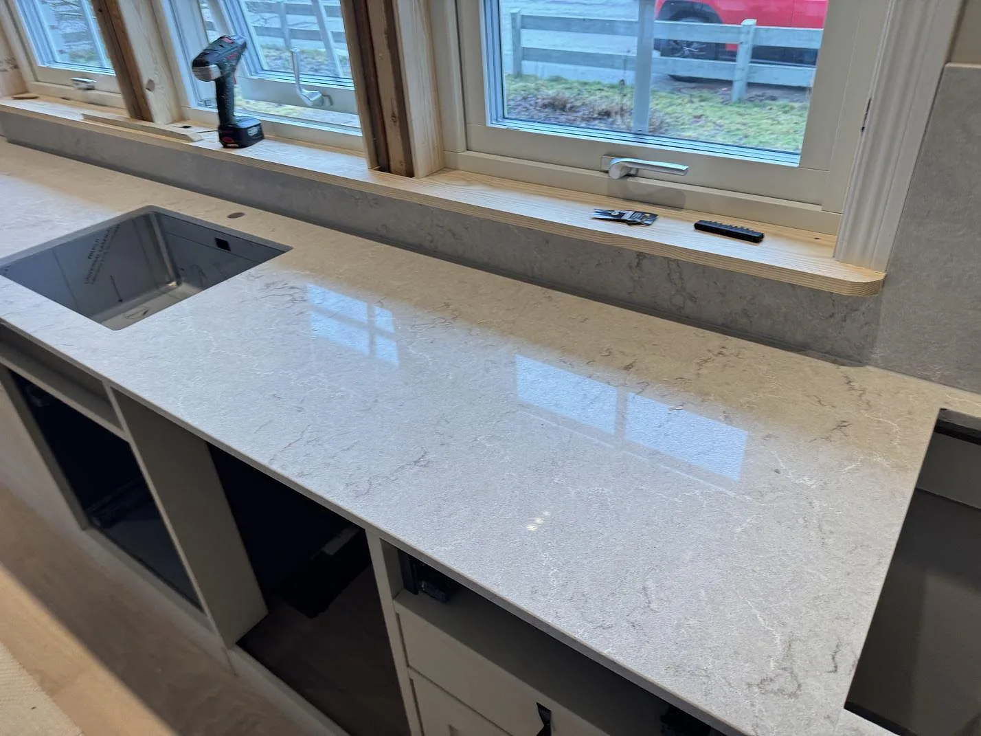 Køkkenbordplade i hvid Caesarstone Bianco Drift, poleret kvartskomposit med grå åretegning, med undermonterad rostfri vask längs fönsterraden i ett køkken i Kalmar.