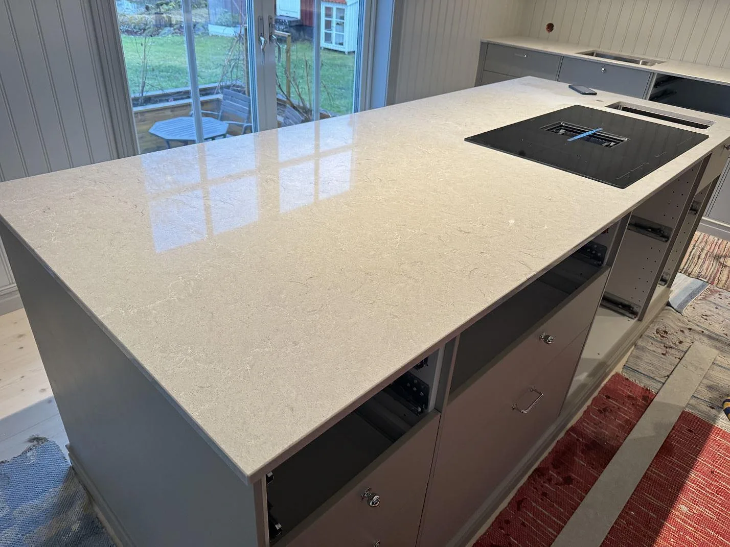 Køkkenø med poleret Caesarstone Bianco Drift i lys kvartskomposit, kogeplade og synlig väggbänk i ett køkken i Vårgårda.