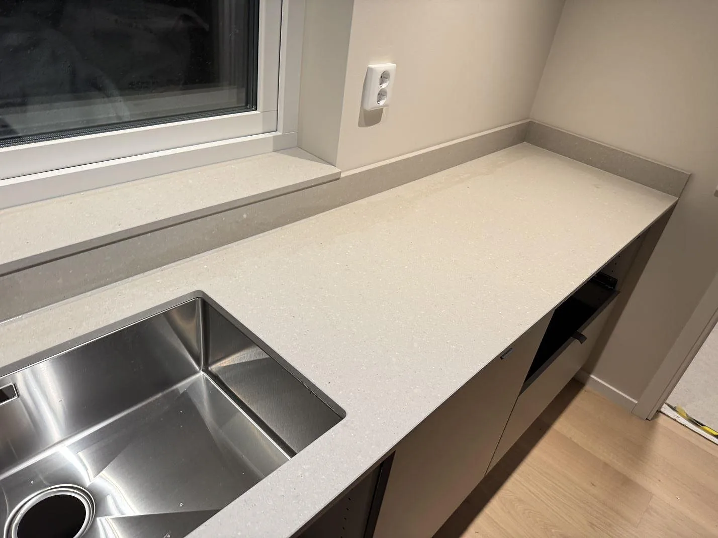 Køkkenbordplade i varm off-white Caesarstone Clamshell, mat kvartskomposit, med underlimmad rostfri vask og vindueskarm i samme sten i ett køkken i Trelleborg.