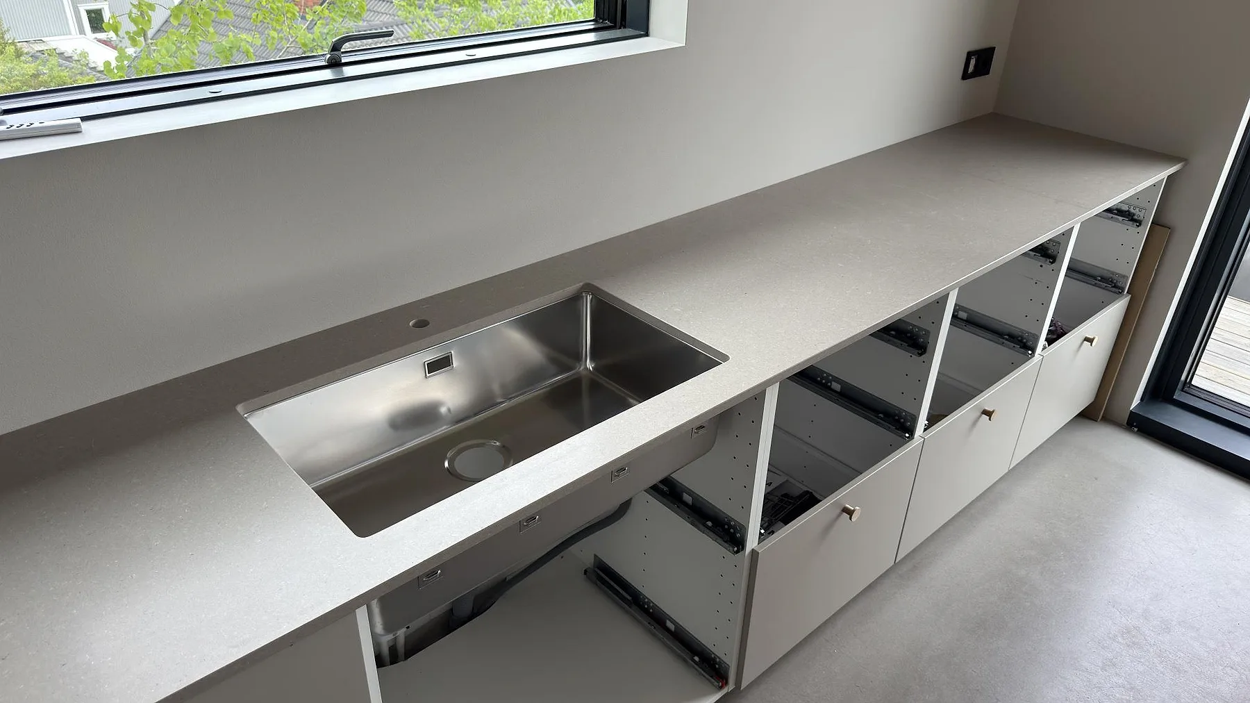Køkkenbordplade i Caesarstone Shitake, en varm beige kvartskomposit med mat overflade, monteret i en lang linje längs fönsterväggen med underlimmad rostfri vask i ett køkken under montering i Saltsjöboo.