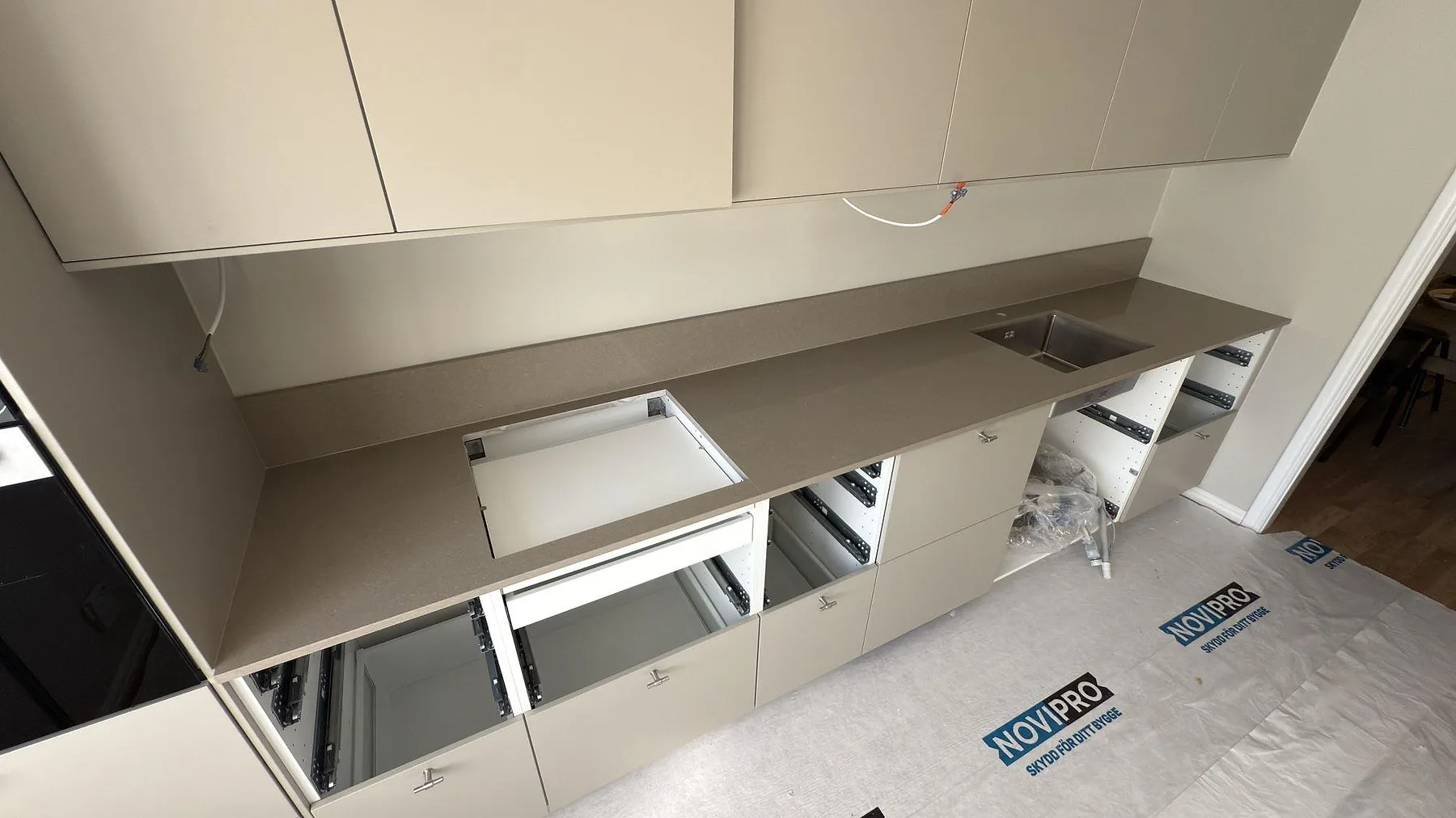 Køkkenbordplade i Caesarstone Shitake, en varm beige kvartskomposit med poleret overflade, monteret i en lang lige linje med underlimmad rostfri vask i ett køkken under montering i Täby.