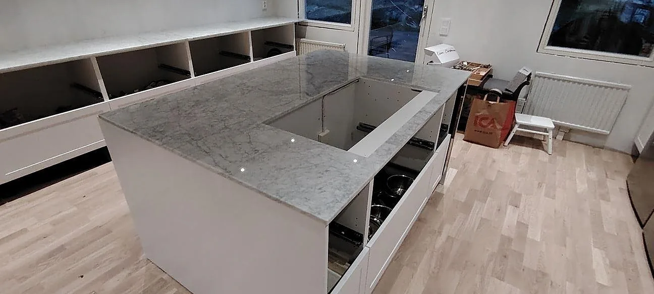 Køkkenø og väggbänk i poleret Carrara C marmor, 20 mm, med synliga udskæring i ett køkken under montering i Åkersberga.