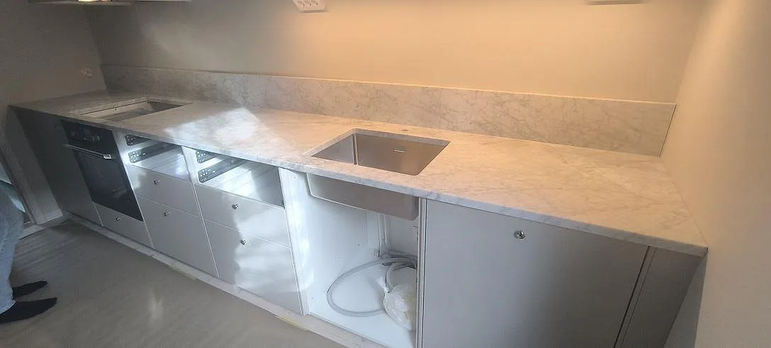 Mat køkkenbordplade i Carrara C marmor med uppvikt stänkkant, synlig vask og hällzon i ett køkken i Älvsjö.