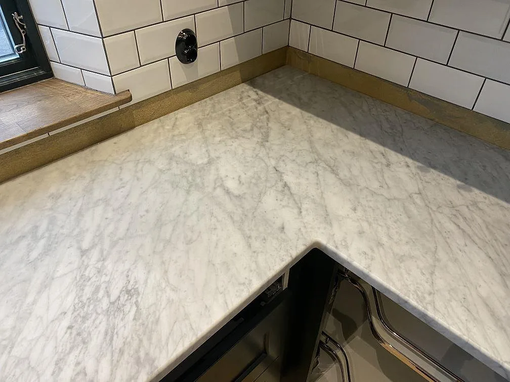 Køkkenbordplade i mat Carrara C-marmor med grå åretegning i L-format hjørner mot vitt kakel i ett køkken i Brantevik.