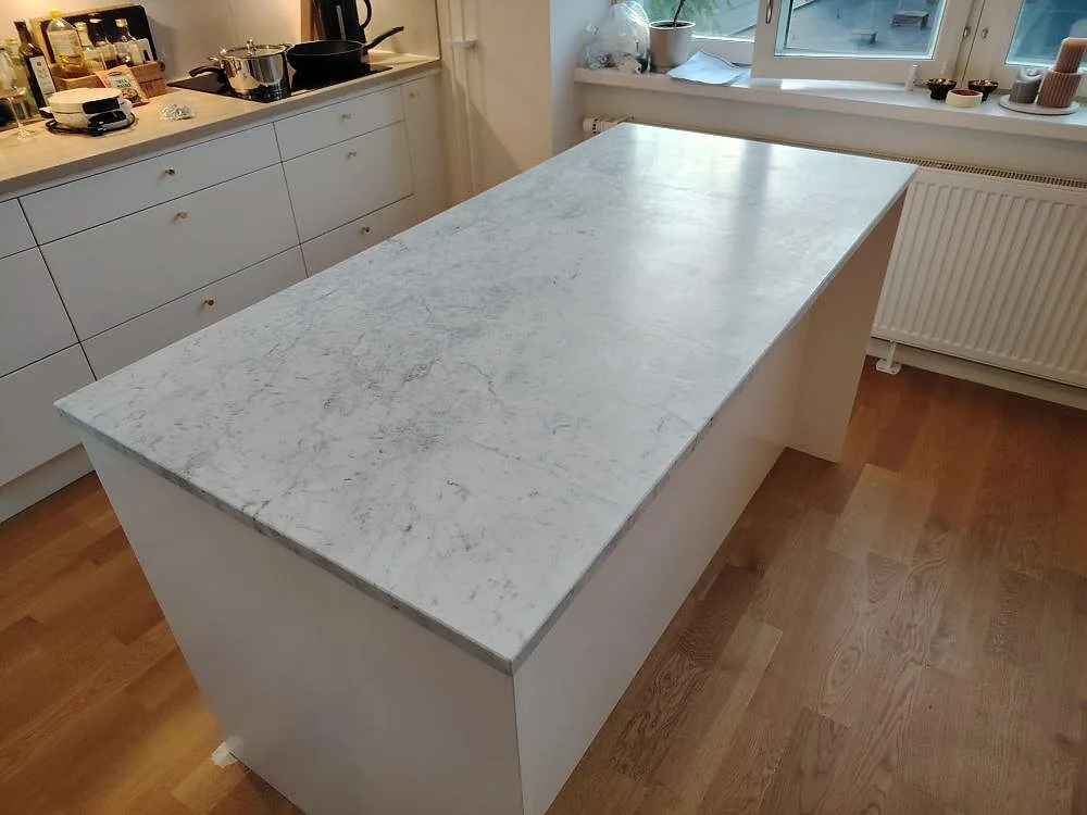 Mat køkkenø i Carrara C marmor, 30 mm, med sammanhängande vitgrå stenyta i ett køkken i Bromma.