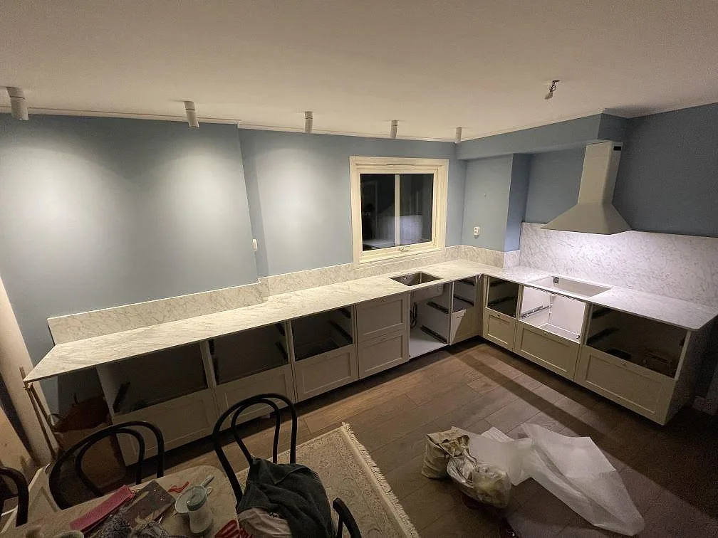 Køkkenbordplade i hvid Carrara C marmor, mat, i ett L-format køkken med duvblå vægge og gräddvita skabe i Bromma, fotograferat under montering.