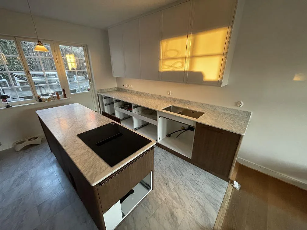 Køkkenbordplade i hvid Carrara C marmor, mat overflade, med underlimmad vask längs væggen og en fristående køkkenø med induktionshäll i ett køkken i Bromma.