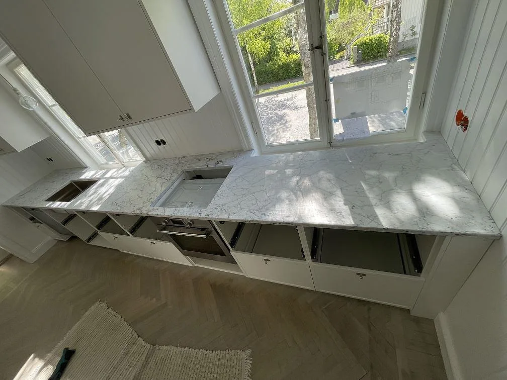 Køkkenbordplade i hvid Carrara C marmor, mat, med grå åretegning og underlimmad rostfri vask, monteret utmed fönstervägg i ett køkken i Bromma.