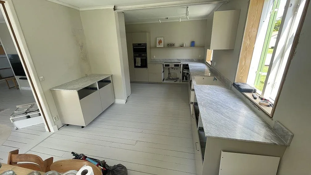Översikt over køkken i Diö med mat køkkenbordplade i Carrara C marmor, uppvikt stänkkant, öppna skabe og fönsterljus.