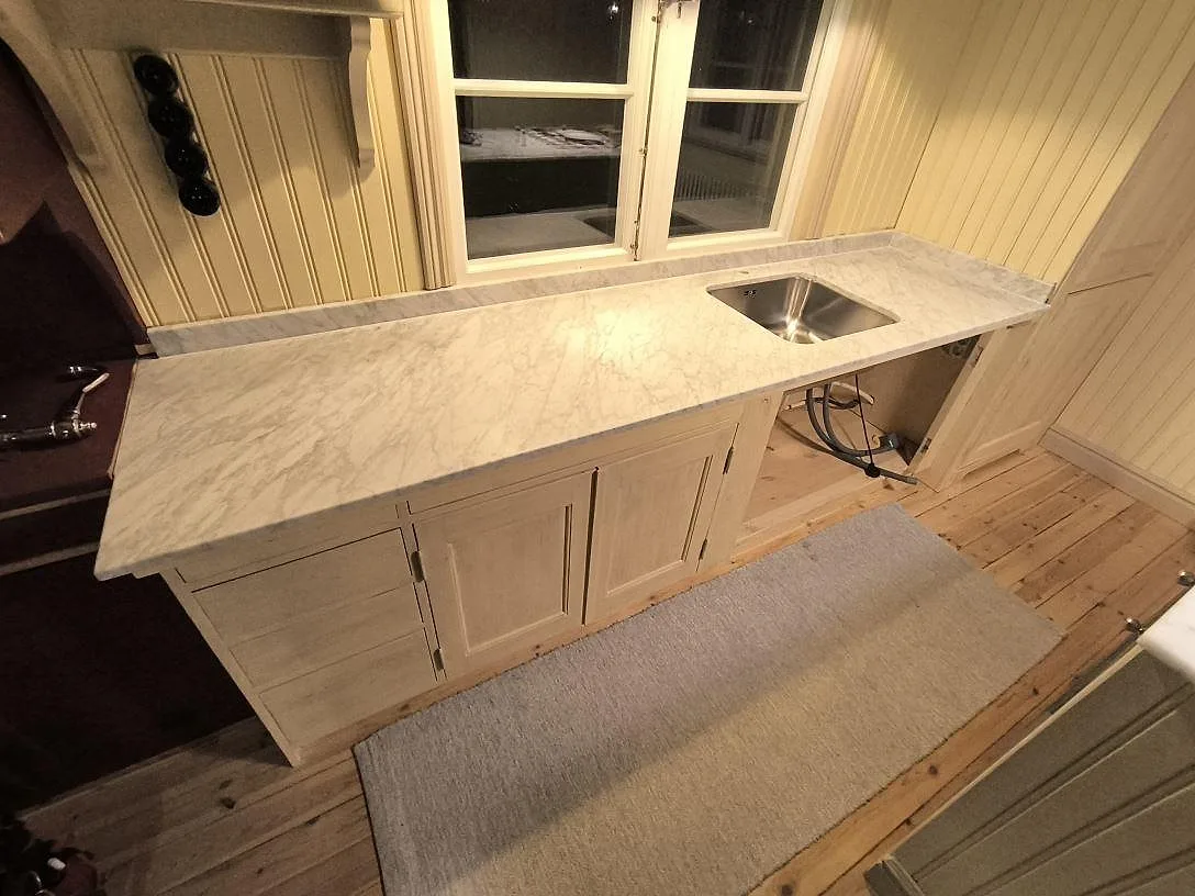 Køkkenbordplade i hvid Carrara C marmor, mat, med låg stänkkant og rostfri vask i ett køkken i Enskede Gård.