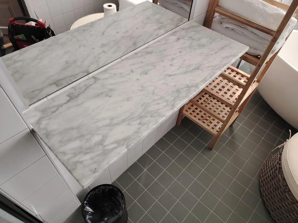 Bordplade i mat Carrara C marmor i ett kaklat badeværelse i Falun, med grå åretegning foran spegel og badkar.