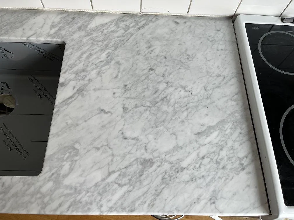 Køkkenbordplade i hvid Carrara-marmor med grå åretegning, poleret, monteret bredvid en sort glaskeramikhäll og hvid kakelvägg i ett køkken i Helsingborg.