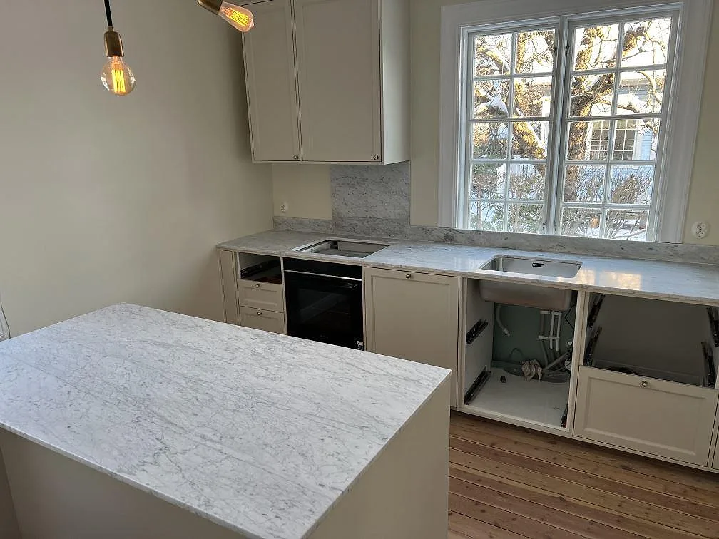 Mat køkkenbordplade i Carrara C marmor, 20 mm, med matchande stænkplade og arbetsyta i ett køkken under montering i Lidingö.