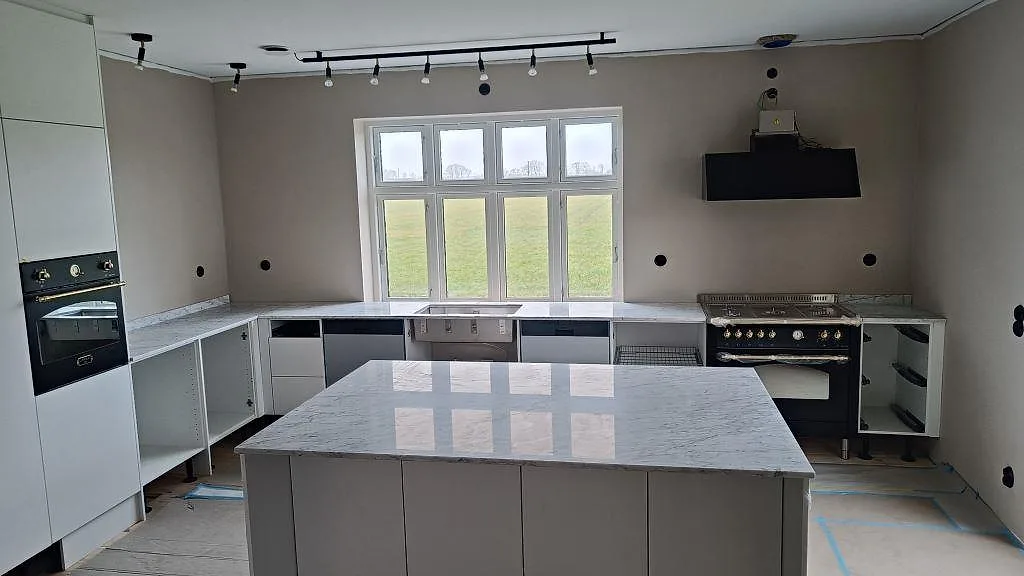 Køkken i Lund med køkkenbordplade og køkkenø i hvid Carrara C marmor, fotograferat under montering med komfur og vindue i bakgrunden.