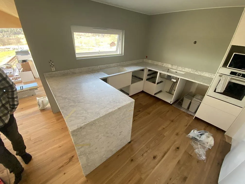 Køkkenbordplade i hvid Carrara C marmor, mat overflade, i ett U-format køkken under montering med vindue og ekgolv i Nygård.