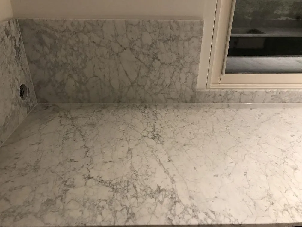 Køkkenbordplade i hvid Carrara C marmor, mat, med vindueskarm og uppvikt stænkplade i samme sten vid fönstret i ett køkken i Sandared.