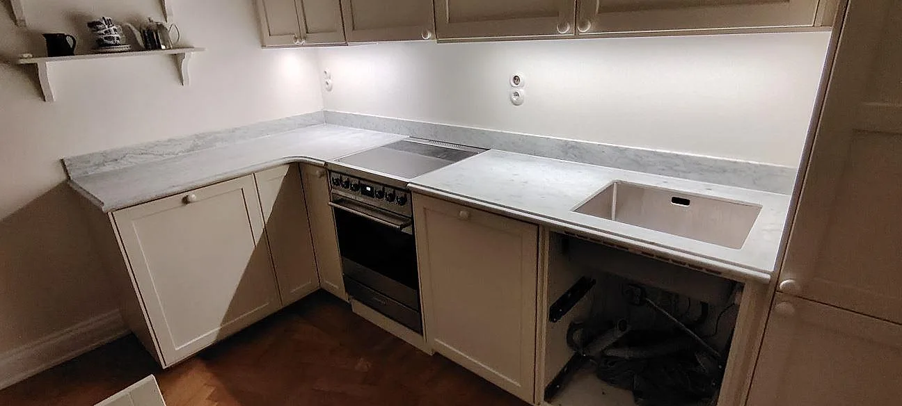 Køkkenbordplade i mat Carrara C marmor med uppvikt stänkkant, underlimmad vask og fristående komfur i ett L-format køkken i Stockholm.