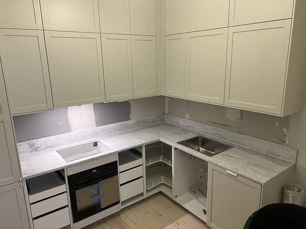 Køkkenbordplade i mat Carrara C marmor med L-form, underlimmad rostfri vask og hällurtag vid ugnen i ett køkken i Stockholm.