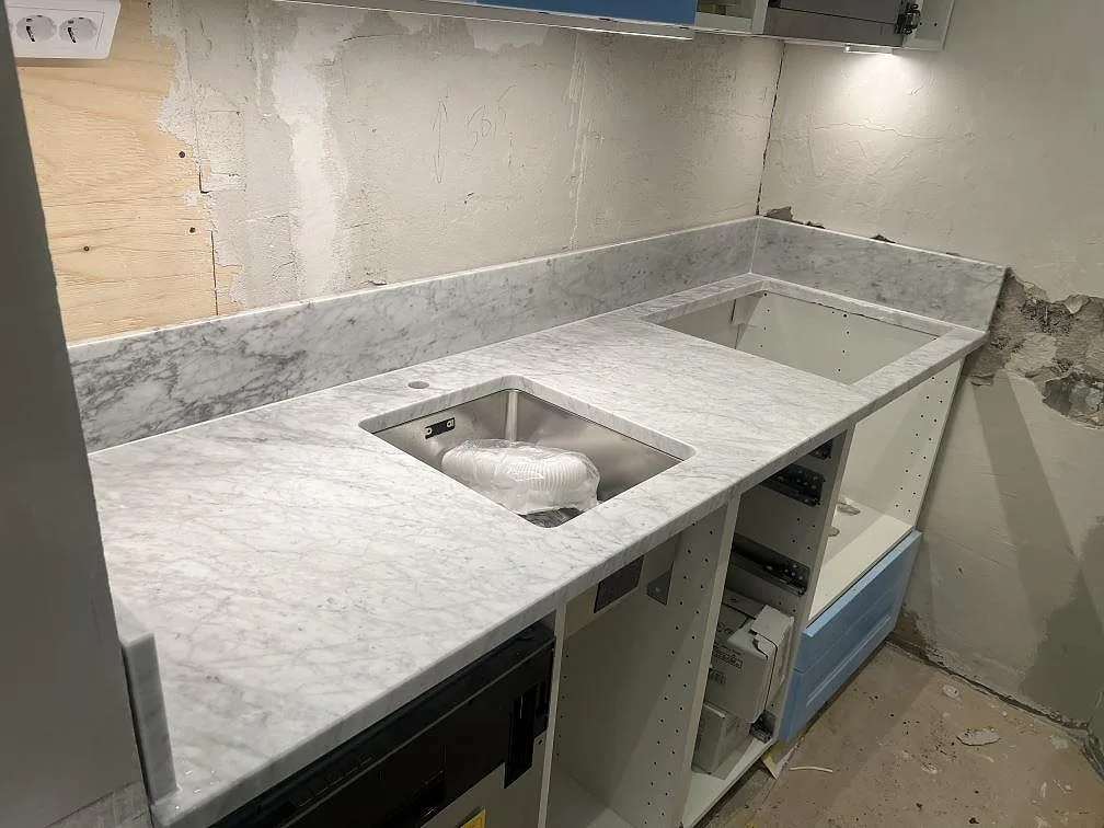 Køkkenbordplade i poleret Carrara C marmor med underlimmad rostfri vask, hällurtag og låg stänkkant i ett køkken i Stockholm.