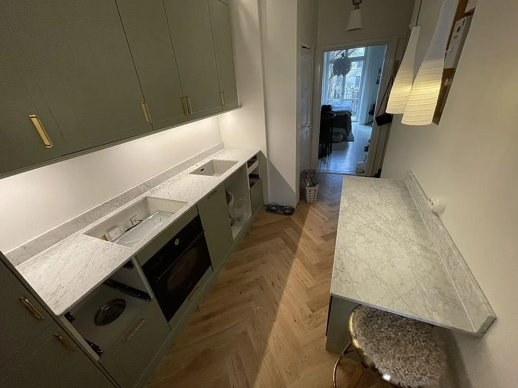 Mat køkkenbordplade i Carrara C marmor med låg stänkkant, diskhoöppning og separat halvö i ett køkken i Stockholm.