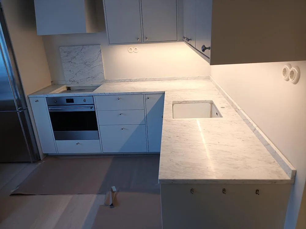 Køkkenbordplade i hvid Carrara C-marmor med grå åretegning, mat overflade, med underlimmad vask og uppvikt stænkplade i ett L-format køkken i Täby.