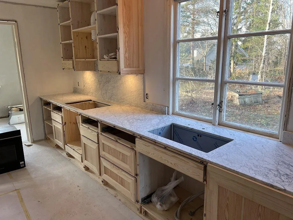 Køkkenbordplade i mat Carrara C marmor, 20 mm, med stænkplade og synligt udskæring vid fönstret i ett køkken i Täby.