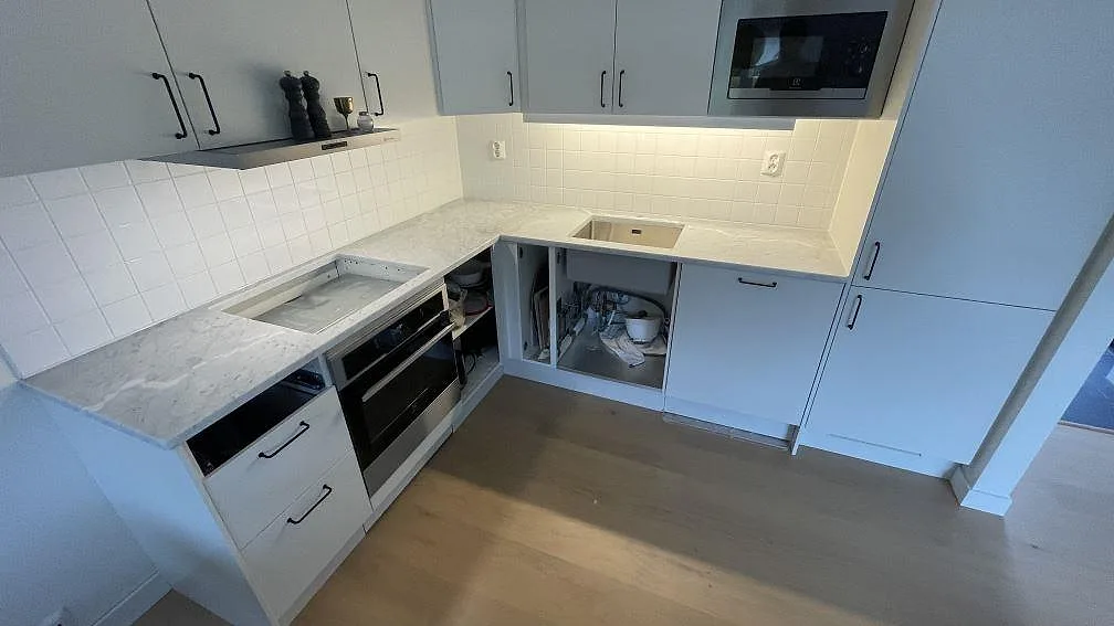 L-formad køkkenbordplade i mat Carrara C-marmor i ett køkken i Torekov, med rostfri vask og plats for kogeplade.
