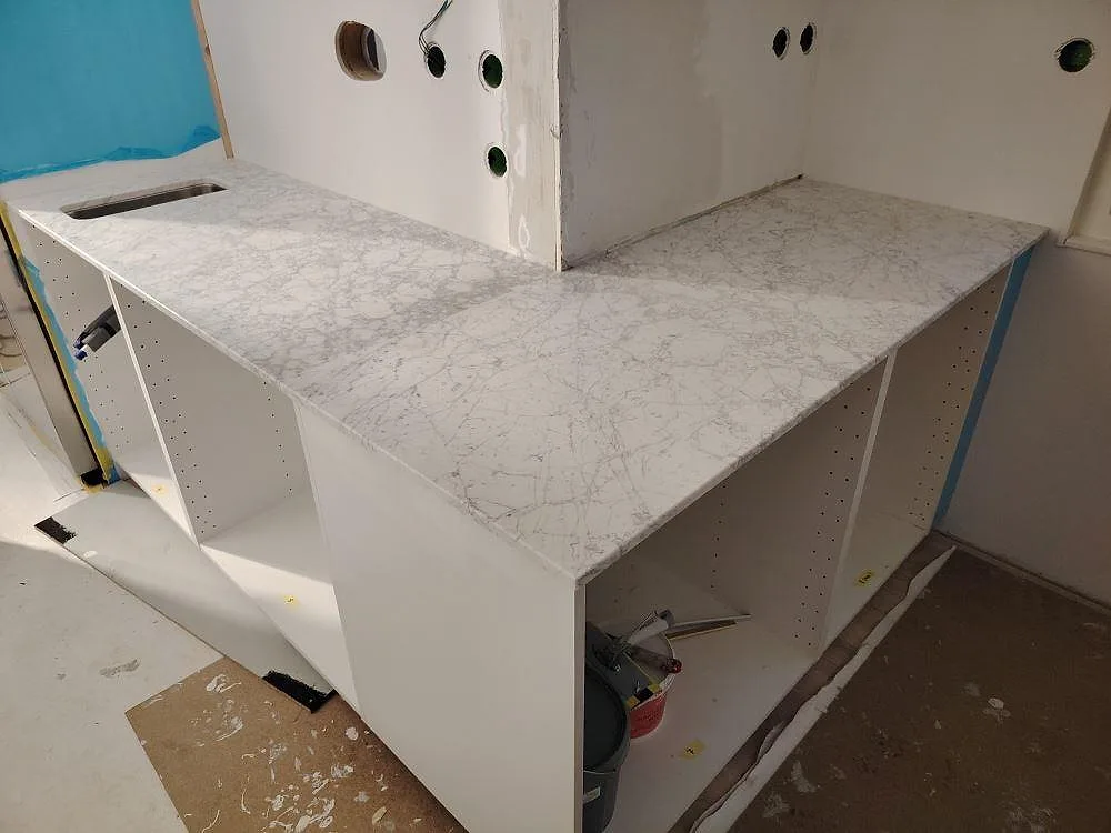 Køkkenbordplade i Carrara C marmor, mat 20 mm overflade, med sammanhängande bordplade rundt om öppna vita skabe i ett køkken i Västervik.