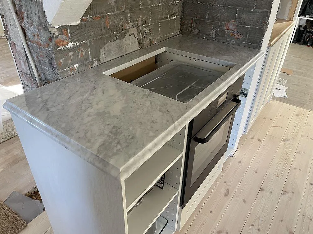 Køkkenbordplade i poleret Carrara C marmor med hällurtag ovanför ovn i ett køkken under montering i Valbo.