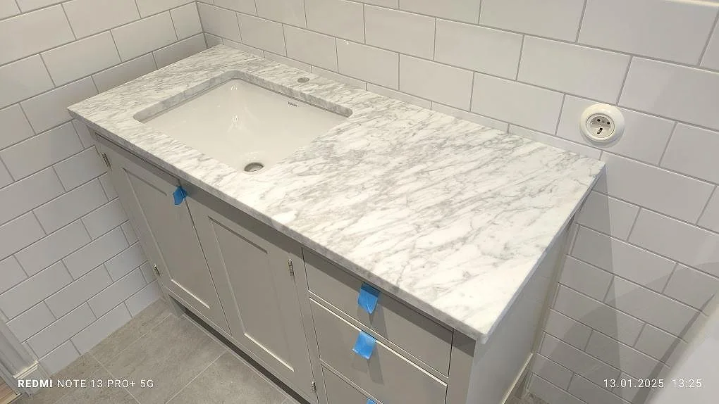 Badrumsbänkskiva i mat Carrara C marmor, 30 mm, med vitt tvättställ på grå kommod mot hvid kakelvägg i Vaxholm.