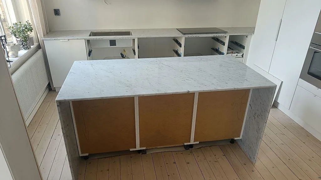 Køkkenbordplade og køkkenø i hvid Carrara C marmor, poleret, med induktionshäll og underlimmad vask i ett køkken under montering i Helsingborg.