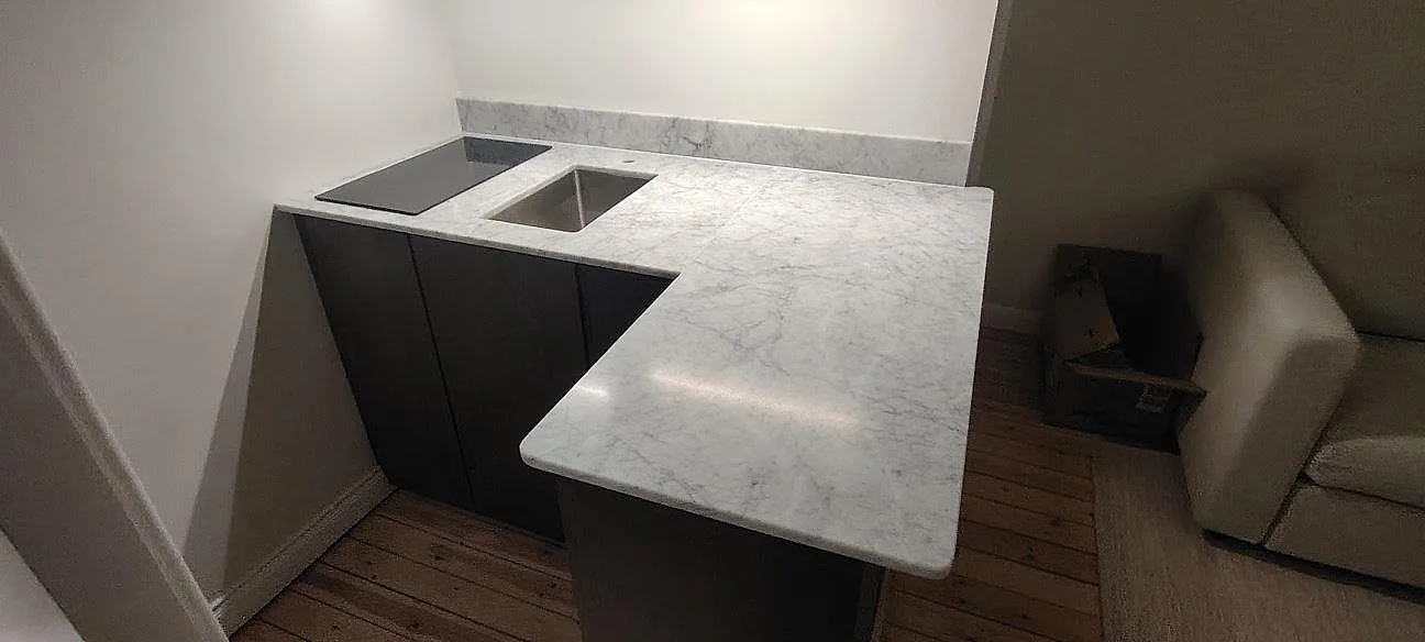 L-formad køkkenbordplade i mat Carrara C marmor med diskhoöppning, kogeplade og låg stänkkant i ett køkken i Stockholm.
