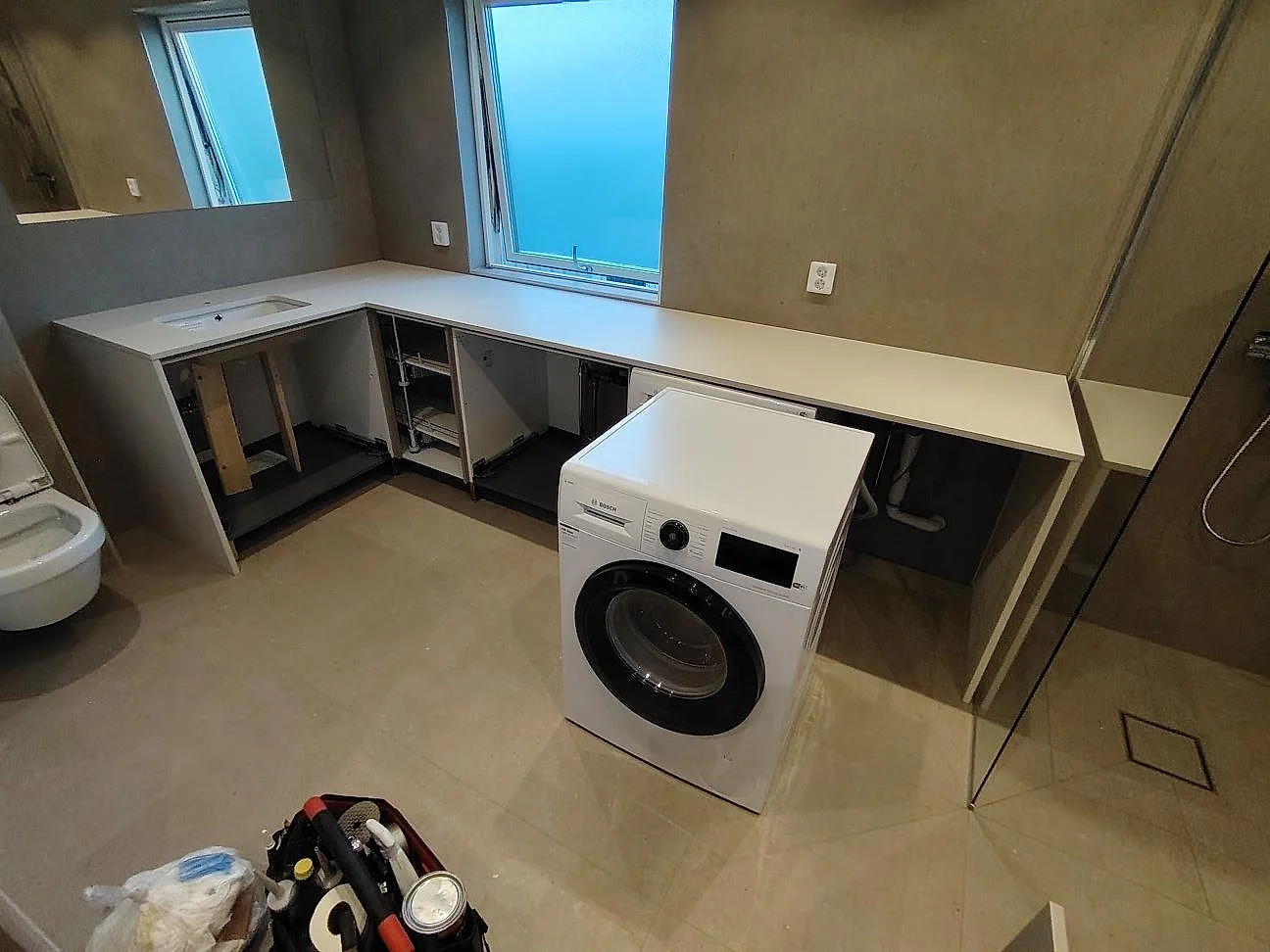 L-formad badrumsbänkskiva i lys mat Dekton Aeris med öppning vid tvättställsplatsen og tvättmaskin i ett badeværelse i Norrtälje.