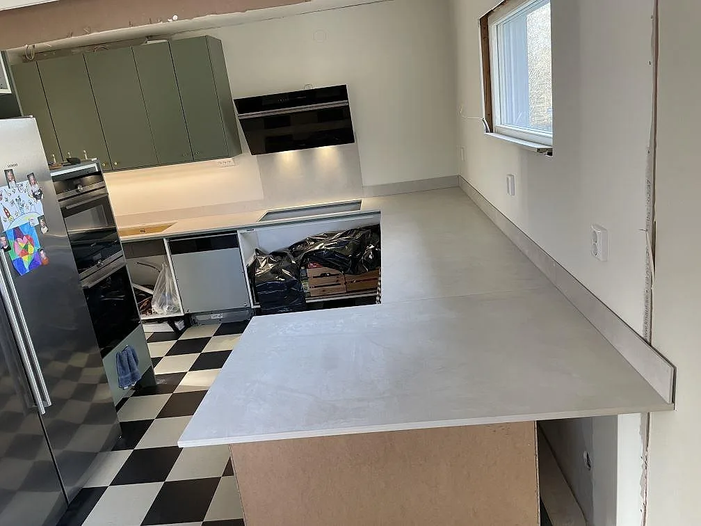 Køkkenbordplade og halvö i ljusgrå mat Dekton Albarium i keramik med kogeplade og låg stänkkant i ett køkken i Vikingstad.