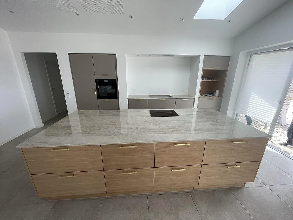 Køkkenbordplade i Dekton Arga, poleret gråbeige keramik, på en stor køkkenø med ekfronter og mässingshandtag i ett nybyggt køkken i Agedrup.