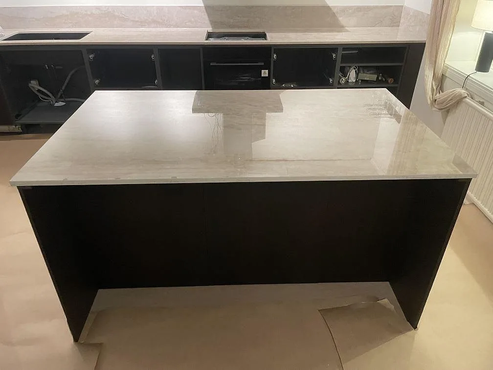 Fristående køkkenø i cremebeige Dekton Arga, poleret keramik med brun-guldig åretegning, foran en sort köksstomme i ett køkken i Flen.