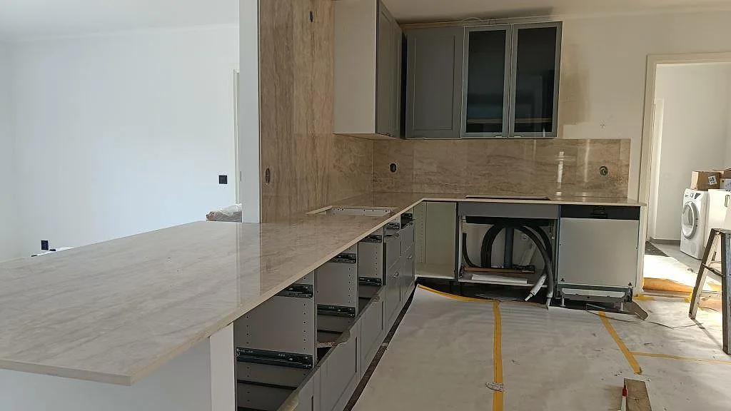 Køkkenbordplade og køkkenø i poleret beige Dekton Arga i ett køkken i Kärna, fotograferat under montering med öppna skabe og heltäckande stänkvägg i samme sten.