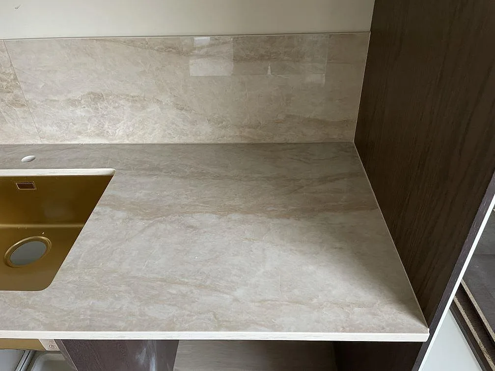 Køkkenbordplade i Dekton Arga, poleret keramik i varm beige med blød åretegning, med uppvikt stænkplade og underlimmad guldfärgad vask i ett køkken i Karlshamn.