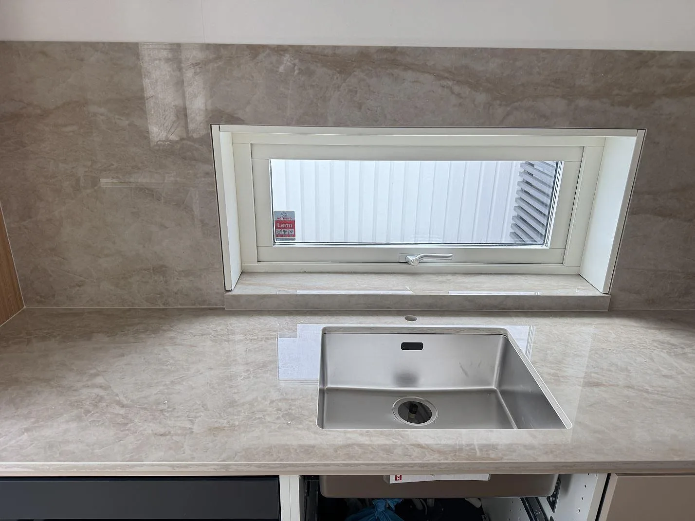 Køkkenbordplade i lys krämbeige Dekton Arga, poleret, med underlimmad rostfri vask og heltäckande stänkvägg i samme keramik vid fönstret i ett køkken i Malmö.