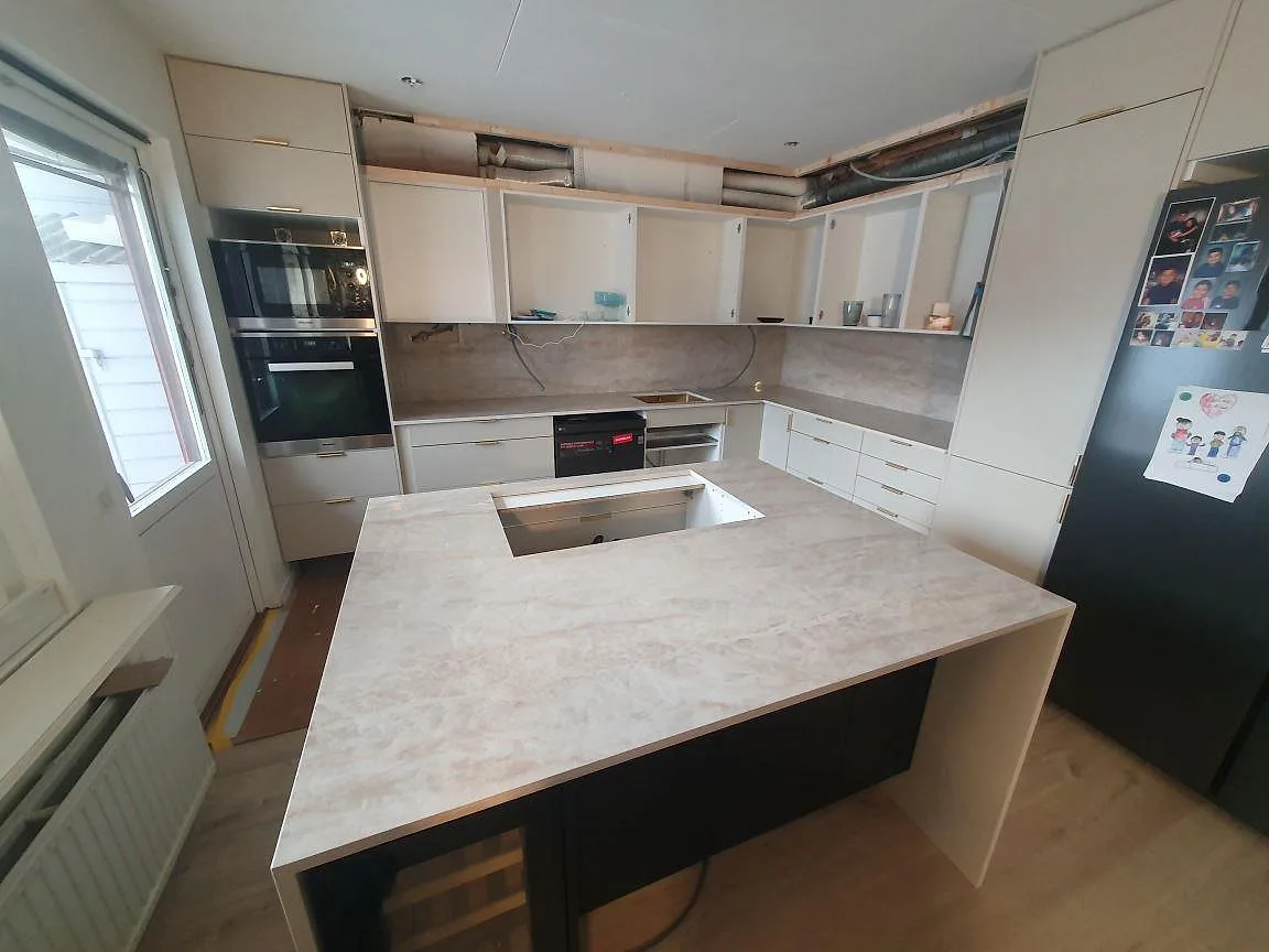 Køkkenbordplade og stor køkkenø i Dekton Arga keramik, varm gräddvit med blød åretegning, poleret, med udskæring for kogeplade i ett køkken under montering i Söderby.