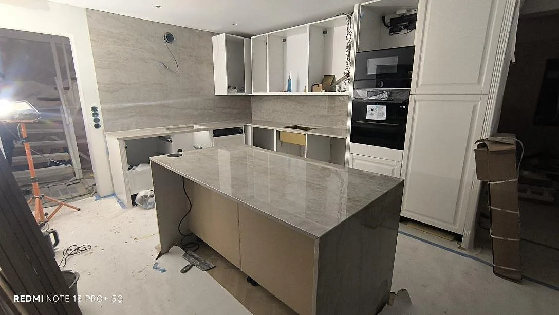 Køkkenø og L-formad køkkenbordplade i poleret Dekton Arga keramik med matchande vægbeklædning i ett køkken under montering i Spånga.