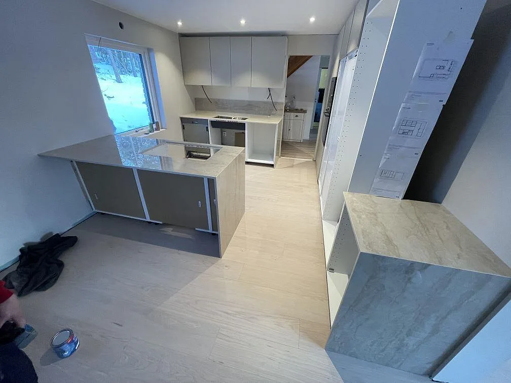 Køkkenbordplade og køkkenø i lys Dekton Arga, poleret, med udskæring for induktionshäll og bänkrad längs væggen i ett køkken under montering i Tyresö.