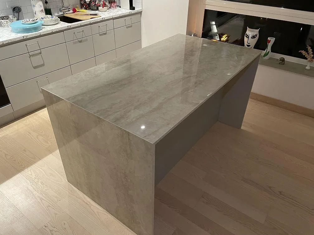 Fristående køkkenø i cremebeige Dekton Arga, 20 mm poleret, med nedvikt stensida mot trägolv og en lys marmorlik bänkrad i bakgrunden i ett køkken i Västervik.