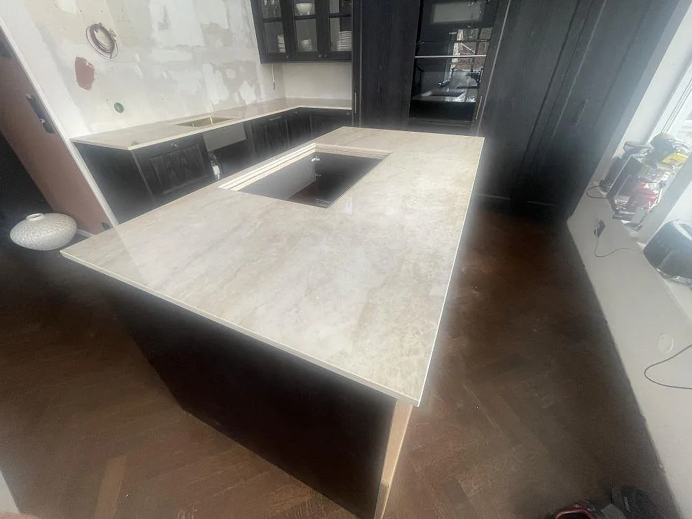 Køkkenbordplade og køkkenø i lys poleret Dekton Arga, keramik, monteret i ett U-format køkken med mørk inredning i Vendelsö.